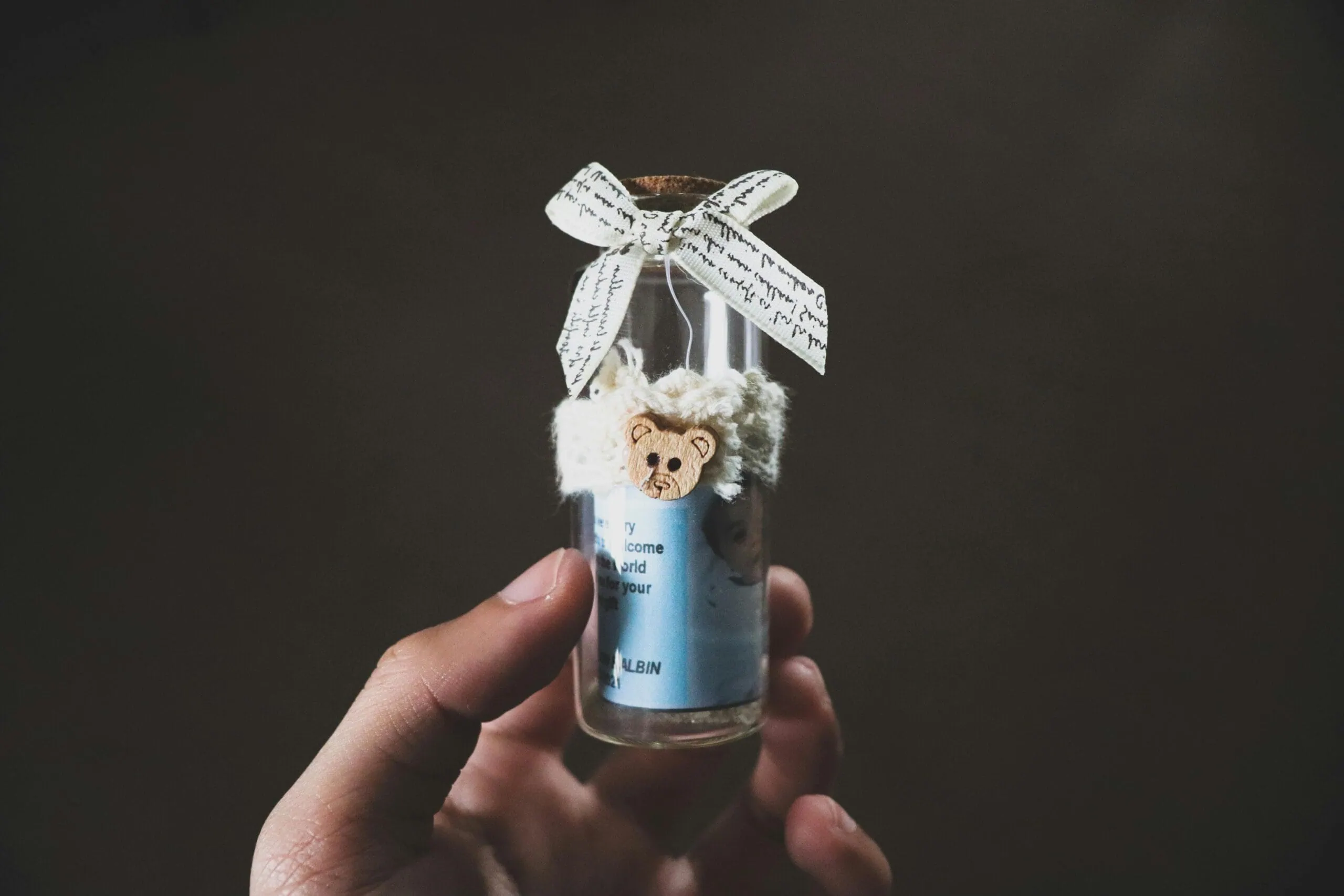 A hand holding a small, decorated glass bottle with a bow and teddy bear embellishment on dark background.